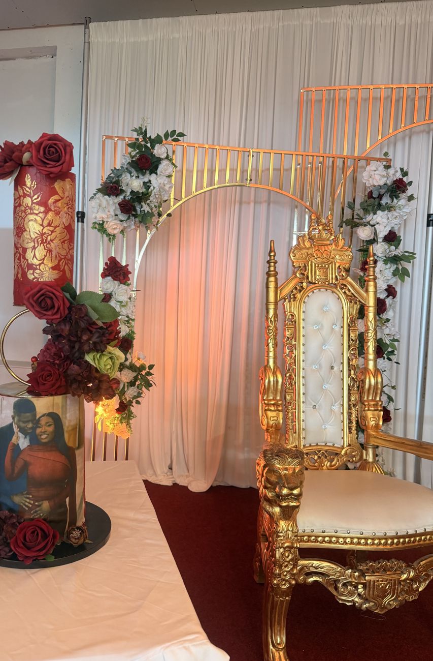Event setup with a decorated cake, flowers, a gold arch, and an ornate gold chair